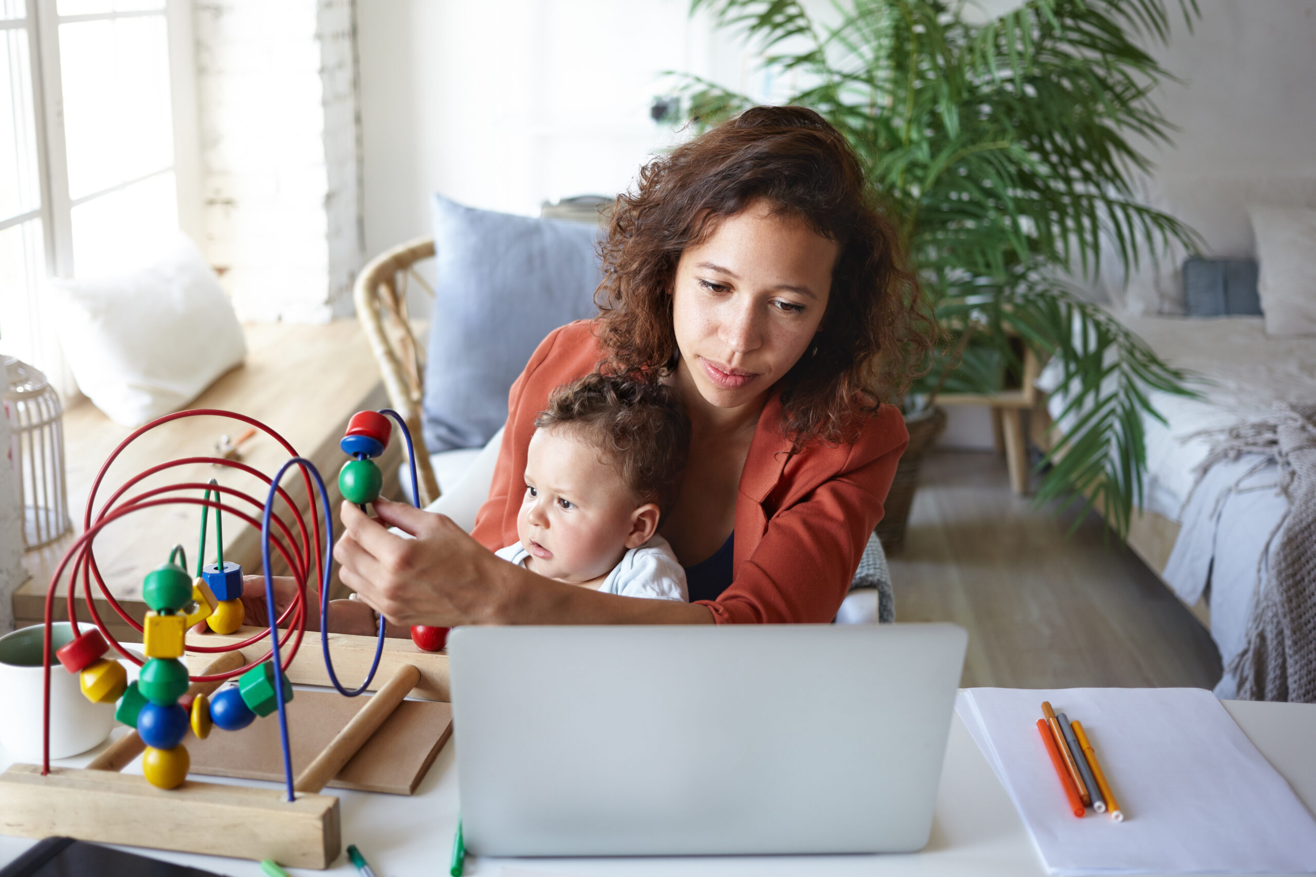 Woman with curly brown hair is sitting and looking at a laptop on a table. A baby sits on her lap and is playing with a bead organizer next to the computer.